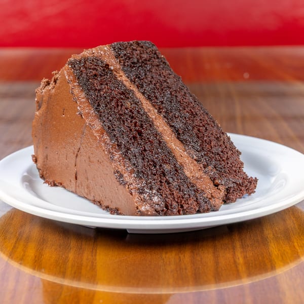 A slice of rich chocolate cake with chocolate frosting on a white plate, on a wooden table.