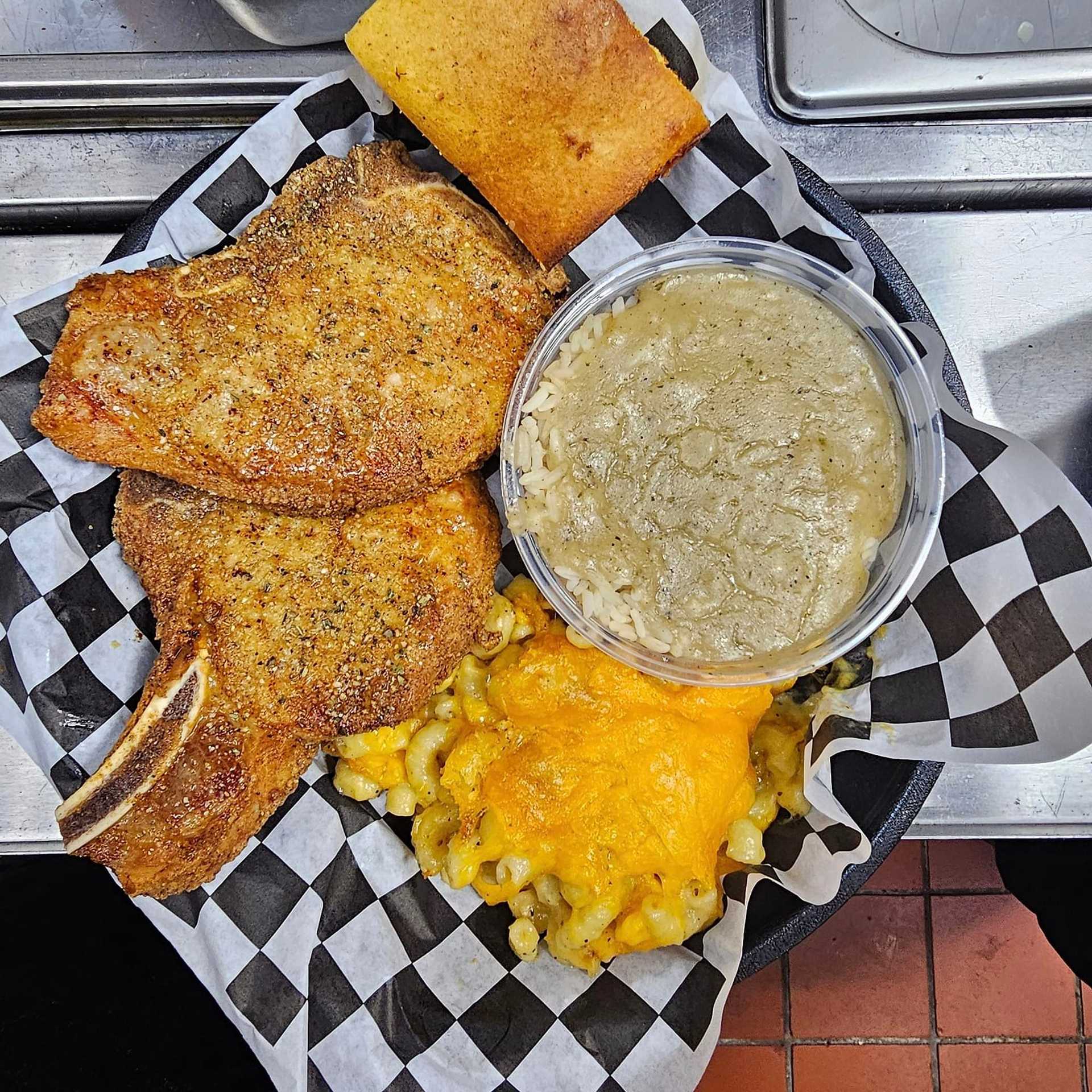 Plate of seasoned pork chops, cornbread, macaroni and cheese, and a side of gravy on a checkered paper.