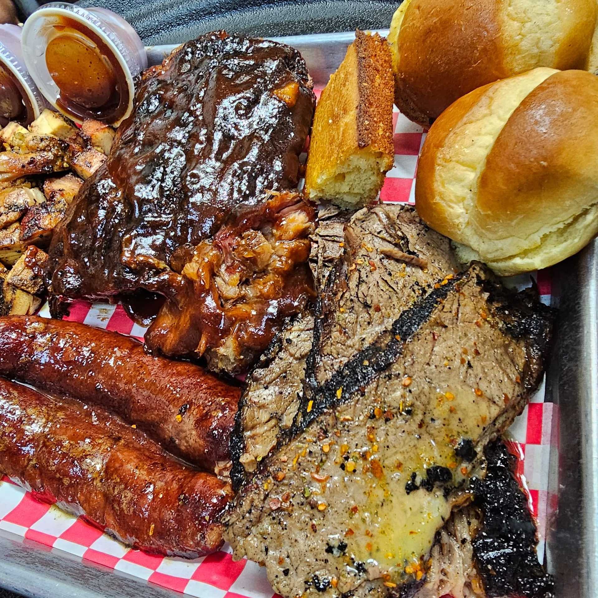 A tray of barbecued meats, including ribs, sausages, brisket slices, and bread rolls with sauces.