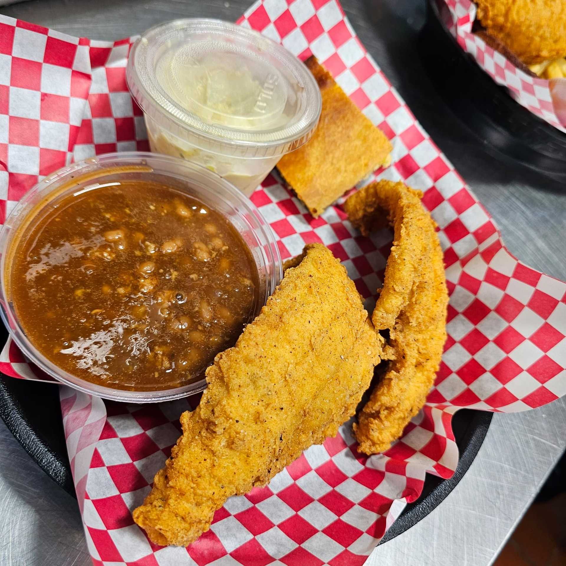 Fried fish with beans, coleslaw, and cornbread in a basket with checkered paper lining.