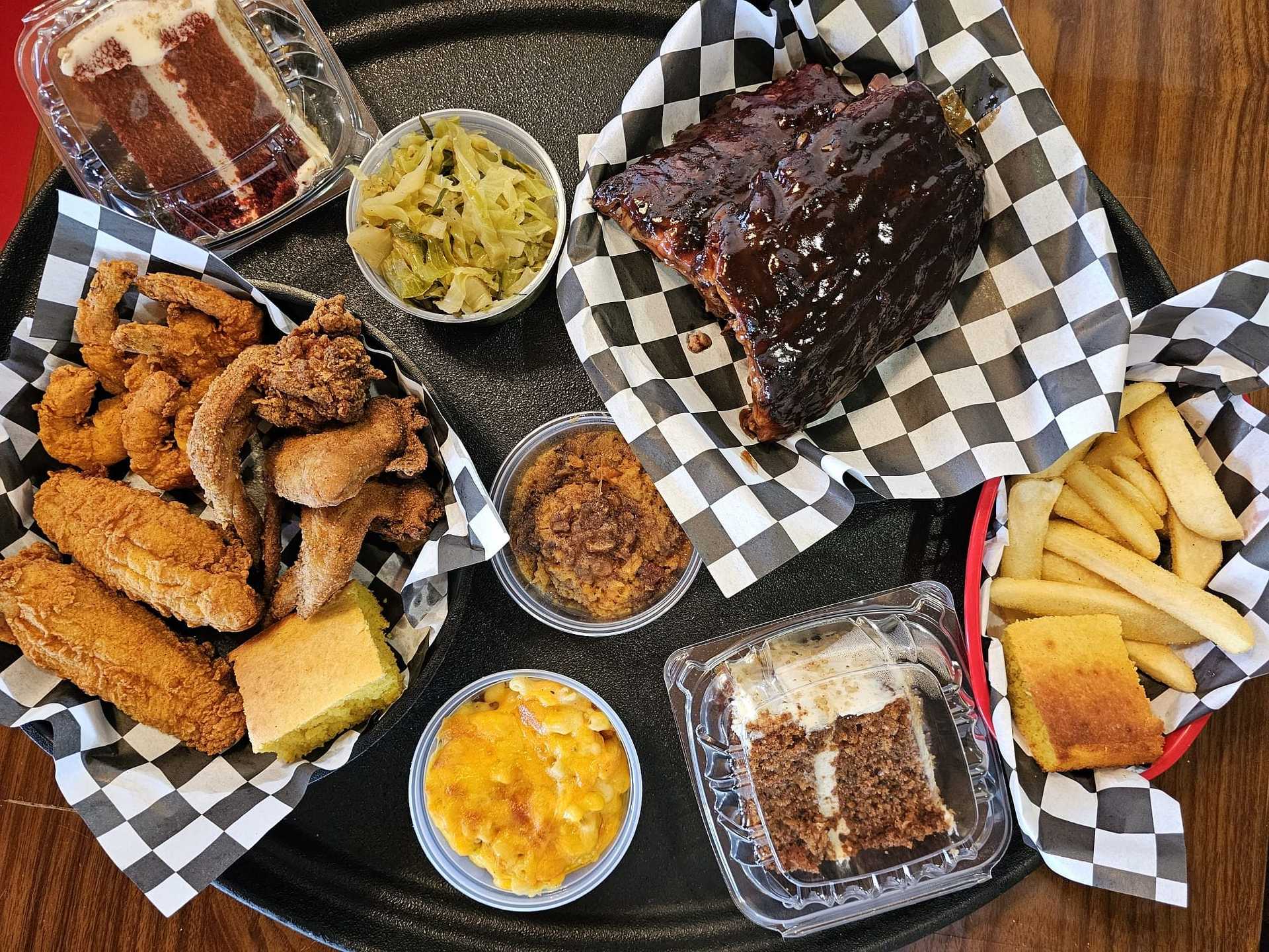 A platter of fried chicken, ribs, fries, cornbread, cake, mac and cheese, and cabbage on checkered paper.