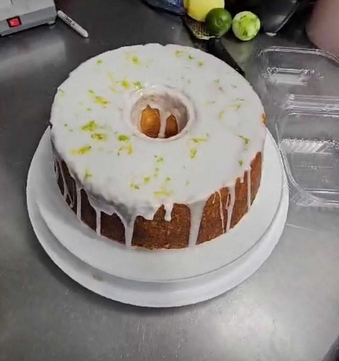 Bundt cake with white glaze and lime zest on a white plate, set on a stainless steel counter.